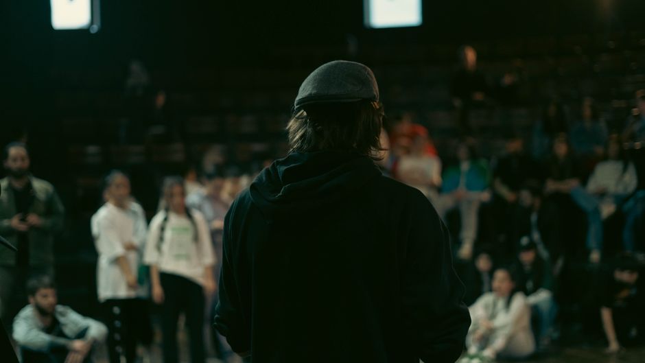 A man in a hoodie and hat stands on stage, facing a crowd during an indoor event.